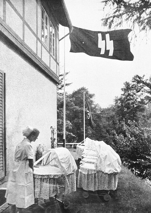 Schwarz-Weiß-Aufnahme einer Krankenschwester mit Kittel und Haube, die in einen von etwa 10 vor einem großen Gebäude aufgereihten Kinderwägen blickt; NSDAP- und SS-Flaggen an Fahnenstangen 