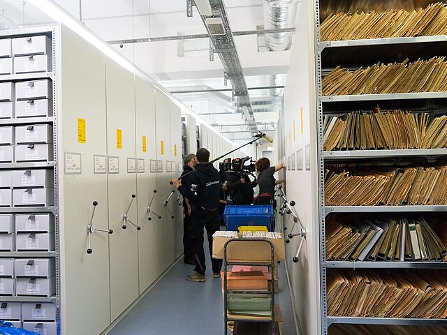 Filmteam im Magazinraum Das Bild zeigt den Blick in einen Gang eines Magazinraumes. Im Vordergurnd sind einige Akten zu erkennen, im Hintergund dreht ein Filmteam.