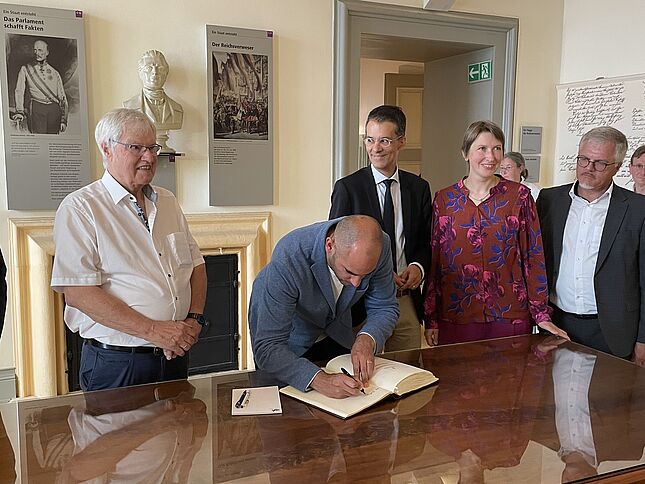 Bild wie sich Dr. Danyal Bayaz (Bildmitte) in das Gästebuch der Bundesarchiv-Erinnerungsstätte Rastatt einträgt