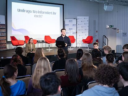 Tag der Pressefreiheit 2026 Eine Bühne ist zu sehen. Vor der Bühne sieht man eine Person stehen und einen Laptop in den Händen halten. Eine Gruppe Jugendlicher sitzt vor ihr auf Stühlen.