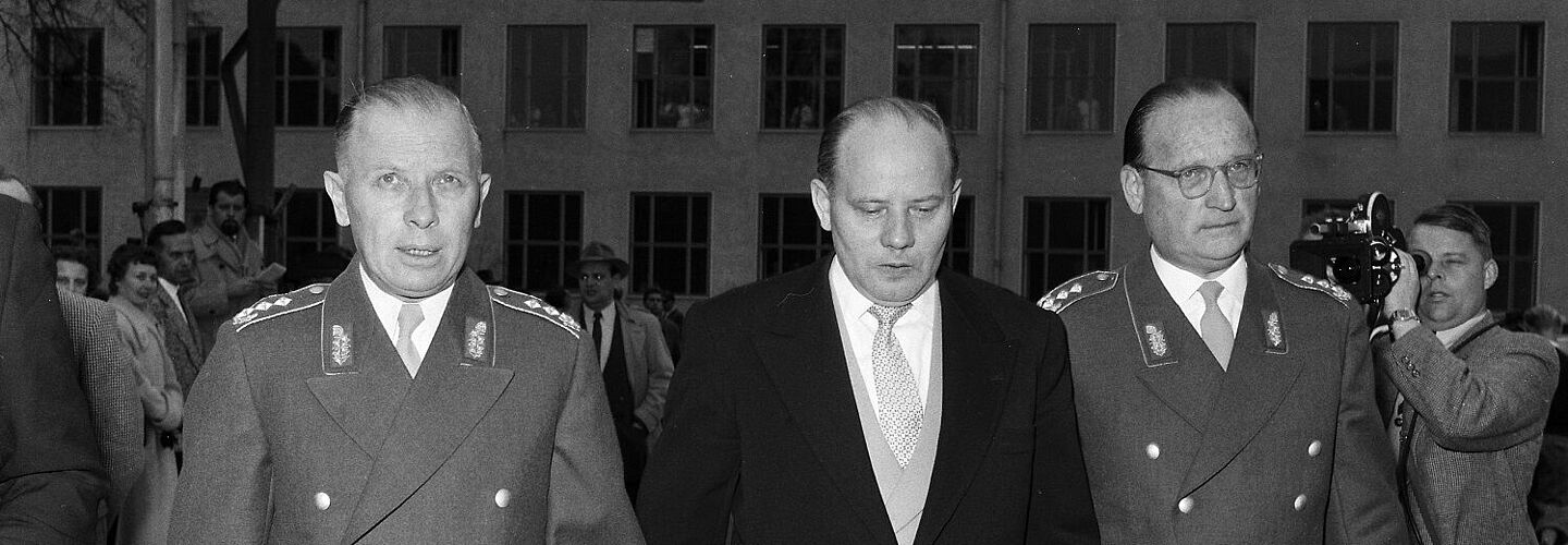 Die Generale Hans Speidel und Adolf Heusinger in Uniform halten ihre Ernennungsurkunden in den Händen. Zwischen ihnen geht Verteidigungsminister Theodor Blank im Anzug. Im Hintergrund sind Presseteams zu erkennen.