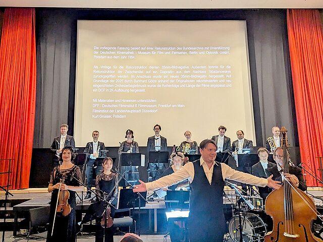 Ein Orchester ist zu sehen. Der Dirigent breitet seine Arme in Richtung der Kamera aus. Im Hintergrund ist eine Kinoleinwand.