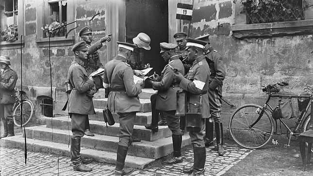 Schwarz-Weiß-Aufnahme mit acht uniformierten Männern, die auf und an einer Treppe vor einem Gebäude stehen und auf Pläne schauen