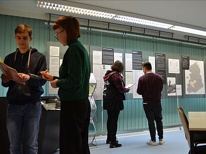 Vier junge Menschen stehen in einem Raum und sehen sich einen Ordner bzw. Ausstellungstafeln an.