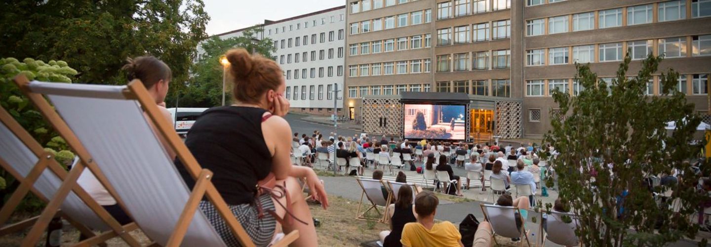 Im Innenhof der ehemaligen Stasi-Zentrale sitzen Menschen, die auf einen großen Kinobildschirm schauen.
