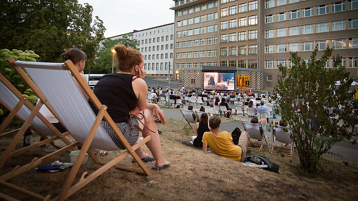 Im Innenhof der ehemaligen Stasi-Zentrale sitzen Menschen, die auf einen groen Kinobildschirm schauen.