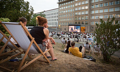 Im Innenhof der ehemaligen Stasi-Zentrale sitzen Menschen, die auf einen großen Kinobildschirm schauen.