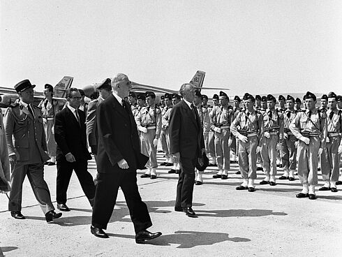 Konrad Adenauer, Charles de Gaulle und weitere Herren gehen entlang einer Soldatentruppe. Im Hintergrund sind zwei Flugzeuge zu sehen.