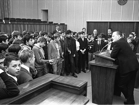 ein im Anzug gekleideter Herr stützt sich auf einem Rednerpult auf und spricht zu einer Gruppe von etwa 50 Jugendlichen, die im Halbkreis um das Pult herum stehen