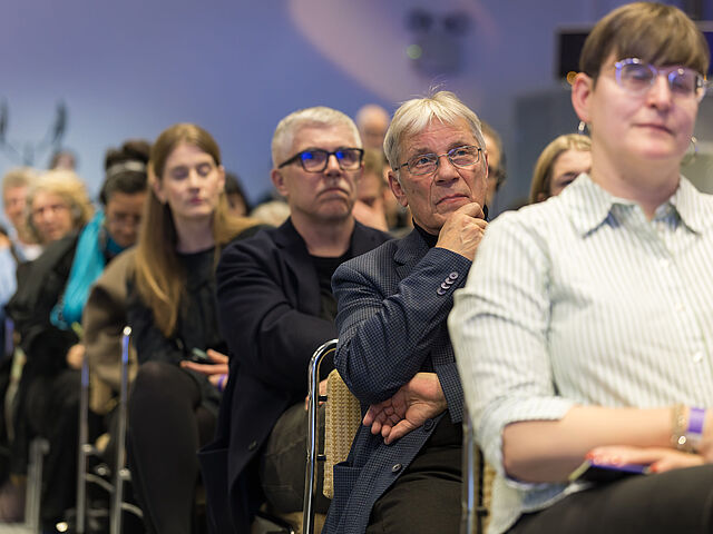 Mehrere Personen sitzen auf Stühlen und schauen konzentriert an der Kamera vorbei.
