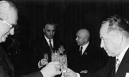 Im Vordergrund stoßen zwie Personen mit Sekt im Glas an. Im Hintergrund sind vier Personen zu erkennen, die ebenfalls ein Sektglas in der Hand haben. Anlass des Bildes war die Verleihung eines Ordens an Erich Mielke.