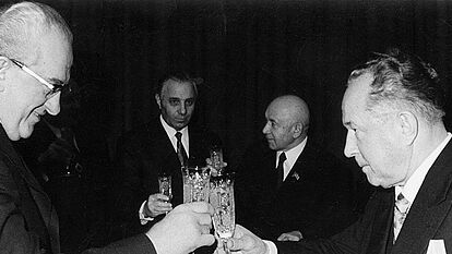 Im Vordergrund stoßen zwie Personen mit Sekt im Glas an. Im Hintergrund sind vier Personen zu erkennen, die ebenfalls ein Sektglas in der Hand haben. Anlass des Bildes war die Verleihung eines Ordens an Erich Mielke.