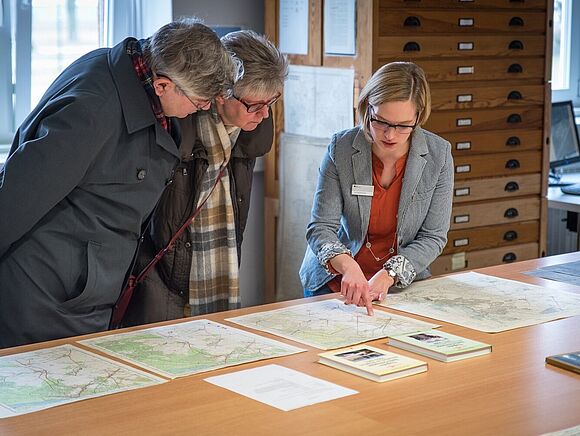 Eine Mitarbeiterin des Bundesarchivs zeigt einer Besucherin und einem Besucher Karten aus den Beständen des Bundesarchivs