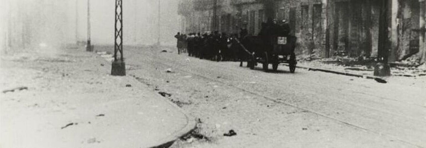Auf dieser Schwarz-Weiß-Fotografie ist eine Straße im Warschauer Ghetto zu sehen. Die Straße ist voller Rauch und Schutt. Die Gebäude sehen stark beschädigt aus. Des Weiteren befinden sich mehrere Menschen in Kolonne laufend auf der Straße. Hinter ihnen fährt eine Kutsche samt Pferd.