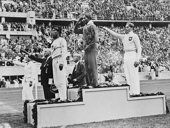 Siegerehrung&nbsp;im Weitsprung bei den Olympischen Spielen 1936 in Berlin. Auf dem Podest&nbsp;(von links nach rechts):&nbsp;Naoto Tajima, Jesse Owens, Carl Ludwig "Luz" Long. Im Hintergrund Theodor Lewald (links neben Tajima)