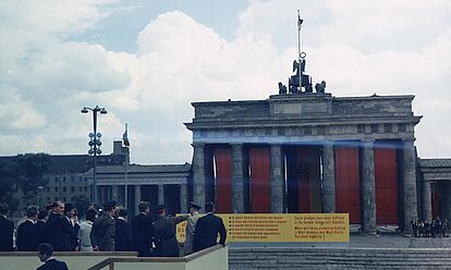 US-Präsident John F. Kennedy steht auf einem Podest und blickt auf das mit roten Tüchern verhängte Brandenburger Tor.