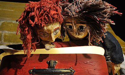 Zwei Handpuppen blicken in einen roten, nicht ganz geöffneten Koffer. Das Innere des Koffers leuchtet leicht.