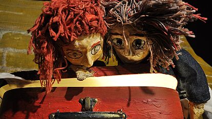 Zwei Handpuppen blicken in einen roten, nicht ganz geöffneten Koffer. Das Innere des Koffers leuchtet leicht.