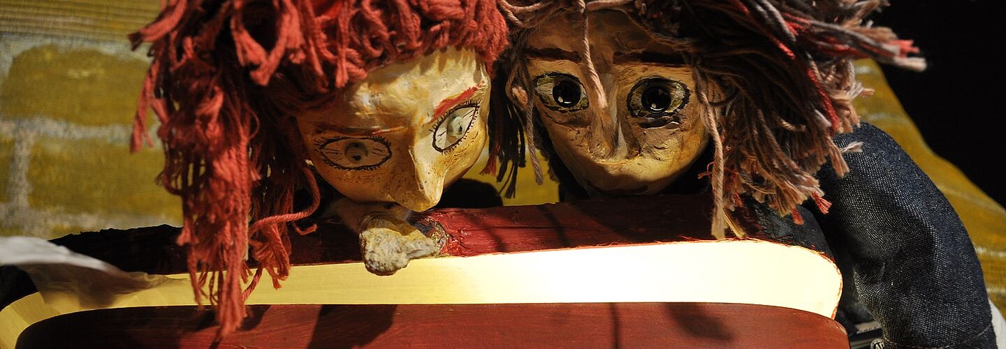 Zwei Handpuppen blicken in einen roten, nicht ganz geöffneten Koffer. Das Innere des Koffers leuchtet leicht.