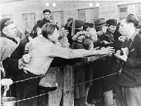 Zivilistinnen und Zivilisten stehen hinter einem Stacheldrahtzaun und strecken ihre Arme in Richtung eines Mannes, der Brot verteilt.