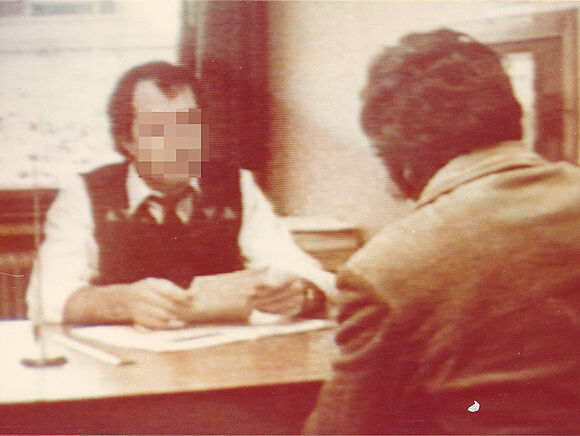 Ein Mitarbeiter des Notaufnahmelagers Gießen sitzt an einem Schreibtisch. Sein Gesicht ist anonymisiert. Er spricht mit einem Mann, der von hinten zu sehen ist.