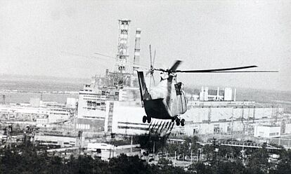 Ein Helikopter versprüht eine Flüssigkeit über einem Waldgebiet. Im Hintergrund ist das verunglückte Kernkraftwerk Tschernobyl zu sehen.