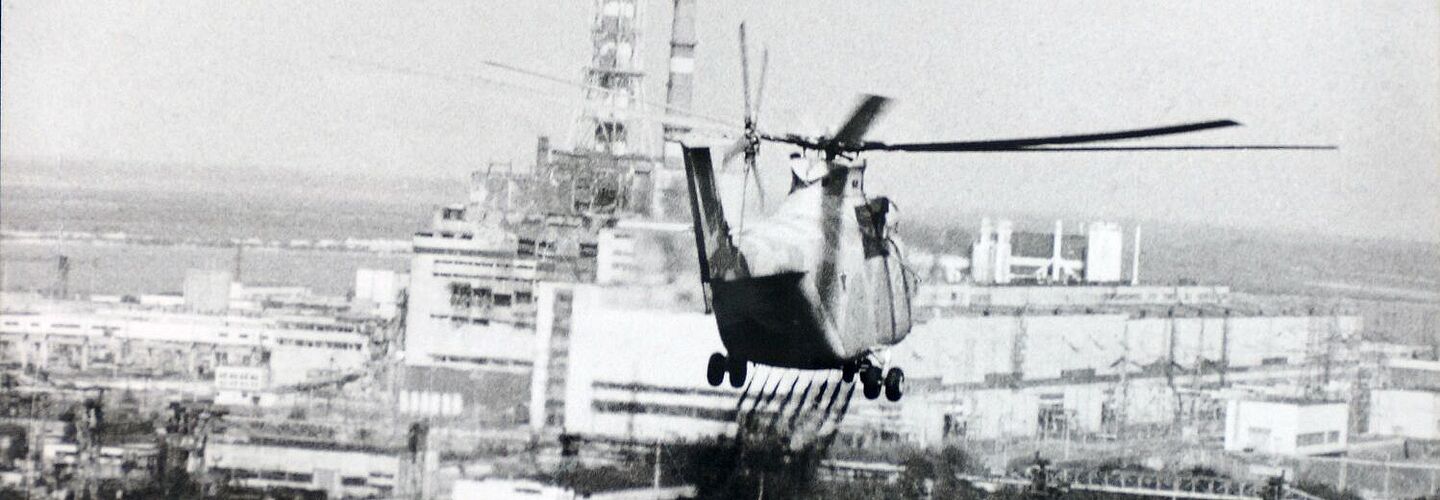 Ein Helikopter versprüht eine Flüssigkeit über einem Waldgebiet. Im Hintergrund ist das verunglückte Kernkraftwerk Tschernobyl zu sehen.