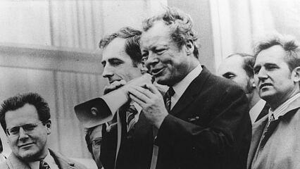 Zu sehen ist eine Gruppe von sechs Männern. Im Vordergrund steht Willy Brandt mit einem Megafon in der Hand, in das er gerade hineinspricht. Am linken Bildrand sieht man Günter Guillaume mit Brille und gemusterter Krawatte.