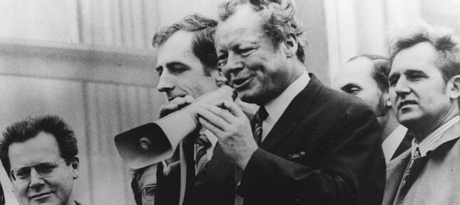 Zu sehen ist eine Gruppe von sechs Männern. Im Vordergrund steht Willy Brandt mit einem Megafon in der Hand, in das er gerade hineinspricht. Am linken Bildrand sieht man Günter Guillaume mit Brille und gemusterter Krawatte.