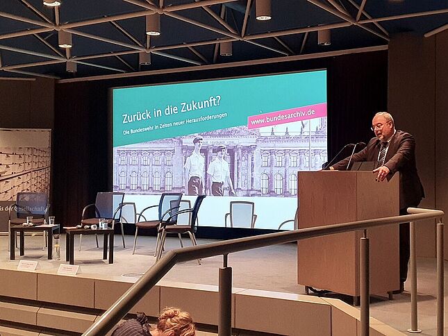 Michael Hollmann bei einer Veranstaltung zum Thema Bundeswehr Ein Mann im Anzug und mit Brille steht an einem Pult auf einer Bühne und spricht ins Mikrofon. Im Hintergrund wird ein Bild projiziert mit den Worten „Zurück in die Zukunft? Die Bundeswehr in Zeiten neuer Herausforderungen“.