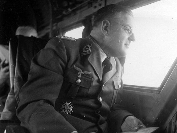 Schwarz-Weiß-Nahaufnahme eines Mannes mit Brille, der eine Karte in den Händen hält und aus dem Fenster eines Flugzeuges schaut. An seiner Uniformjacke hängen mehrere Orden und Abzeichen.