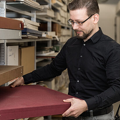 Ein Archivar des Bundesarchivs nimmt Archivalien aus einem Regal eines Magazins., Quelle:
                                                <span class="nanospell-typo">BArch</span>, B 198 Bild-2017-0301-031 / Nobel, Jürgen