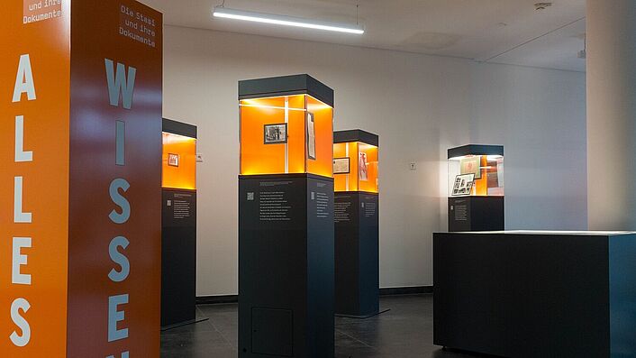 Blick in die Wanderausstellung. Zu sehen sind mehrere Stelen mit Glasvitrinen, in denen sich Dokumente, Fotos und Objekte befinden.