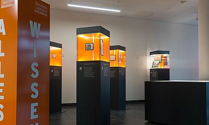 Blick in die Wanderausstellung. Zu sehen sind mehrere Stelen mit Glasvitrinen, in denen sich Dokumente, Fotos und Objekte befinden.