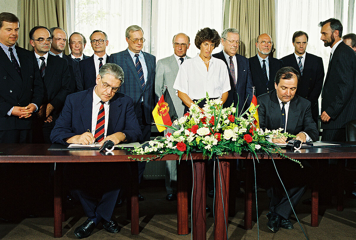 Farbfotografie mit zwei Herren in Anzügen, an einem Tisch sitzend und beim Unterzeichnen von Dokumenten. Auf dem Tisch befinden sich ein Blumenbukett und die Staatsflaggen der DDR und Bundesrepublik Deutschland. Hinter dem Tisch stehen weitere formell gekleidete Personen.