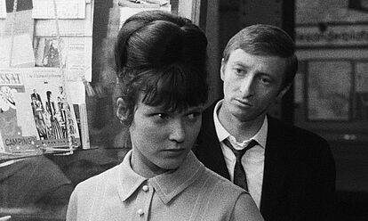 Ein Mann und eine Frau stehen hintereinander vor dem Schaufenster eines Buchladens. Die Frau hat hochgesteckte Haare und wendet ihren Kopf dem hinter ihr stehenden Mann zu. Dieser trägt ein Jacket und eine Krawatte.