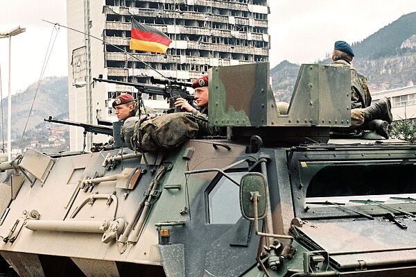 Drei Soldaten der Bundeswehr auf einem Transportpanzer „Fuchs“ in der Innenstadt von Sarajevo. Zwei sind mit Scharfschützengewehren ausgestattet. Im Hintergrund sieht man ein ausgebombtes Hochhaus.