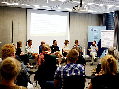 Mehrere Menschen sitzen auf Stühlen und hören einer Person zu, die gerade spricht.