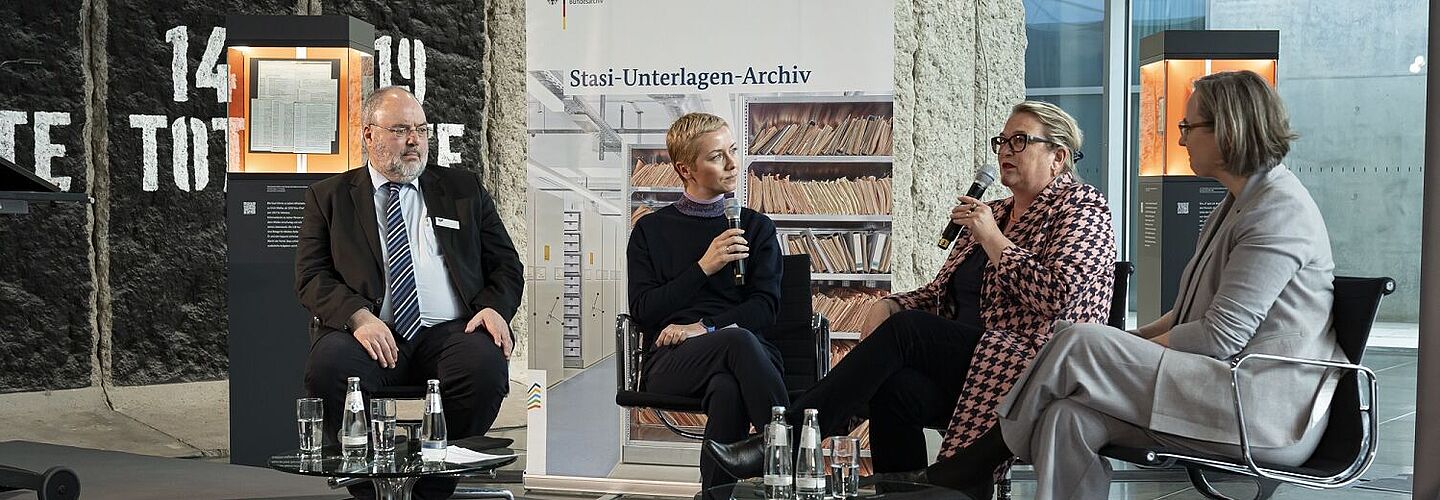 Drei Frauen und ein Mann sitzen auf einer Bühne und sprechen miteinander. Im Hintergrund sind eine Obejktstele und ein Banner mit der Aufschrift 'Stasi-Unterlagen-Archiv' zu sehen.