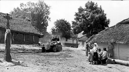 Ein leichter Schützenpanzer der Wehrmacht, in dem drei Soldaten sitzen, fährt über den Platz einer Ortschaft, auf dem ein Kreuz steht. Vor einer Hütte mit Schilf- bzw. Reetdach stehen mehrere Zivilistinnen und Zivilisten.