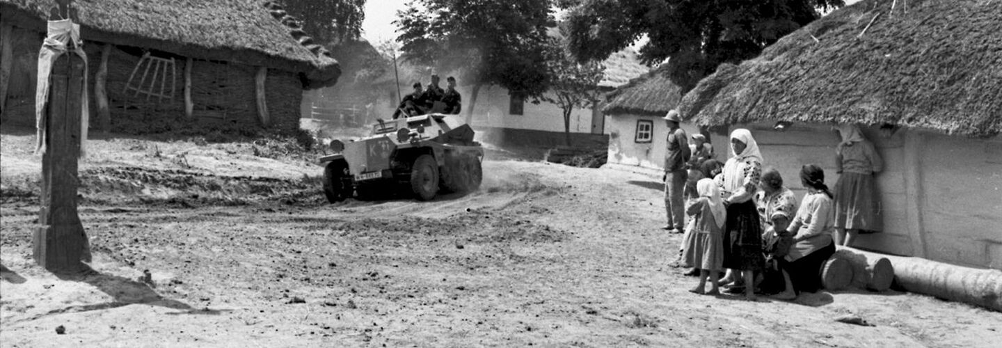 Ein leichter Schützenpanzer der Wehrmacht, in dem drei Soldaten sitzen, fährt über den Platz einer Ortschaft, auf dem ein Kreuz steht. Vor einer Hütte mit Schilf- bzw. Reetdach stehen mehrere Zivilistinnen und Zivilisten.