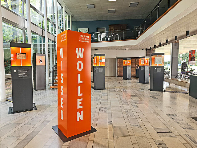 Zu Gast im Staatsarchiv Bremen, 24. September 2026 In einem hohen Saal mit beidseitigen Glassfronten stehen dunkelgraue Ausstellungstelen mit orangenen Vitrinen.