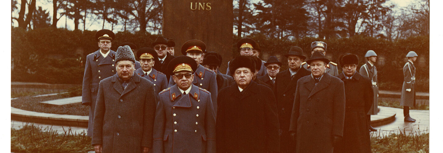 Eine Abordnung von Offizieren der Stasi und des sowjetischen KGB in Uniform und Zivil vor einer Stele auf der steht 'Die Toten mahnen uns'