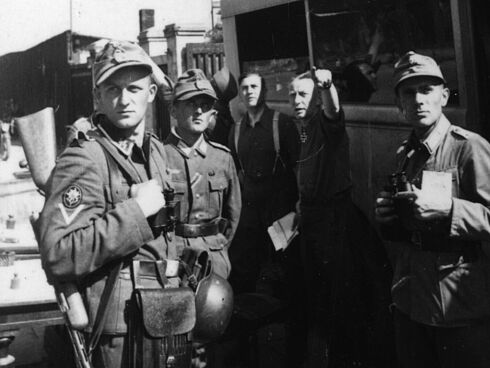 Eine Gruppe wartender Gebirgsjäger, von denen einer mit einer Maschinenpistole bewaffnet ist, steht neben einer Straßenbahn.