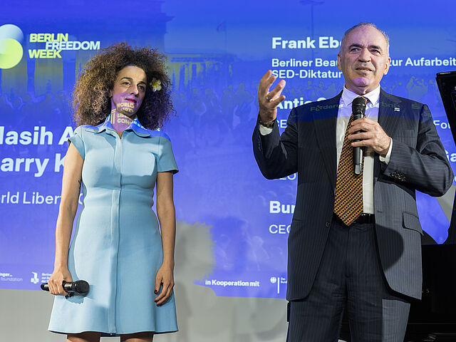 Zwei Personen mit Mikrofon sind zu sehen. Die rechte Person trägt einen Anzug und spricht ins Mikrofon.