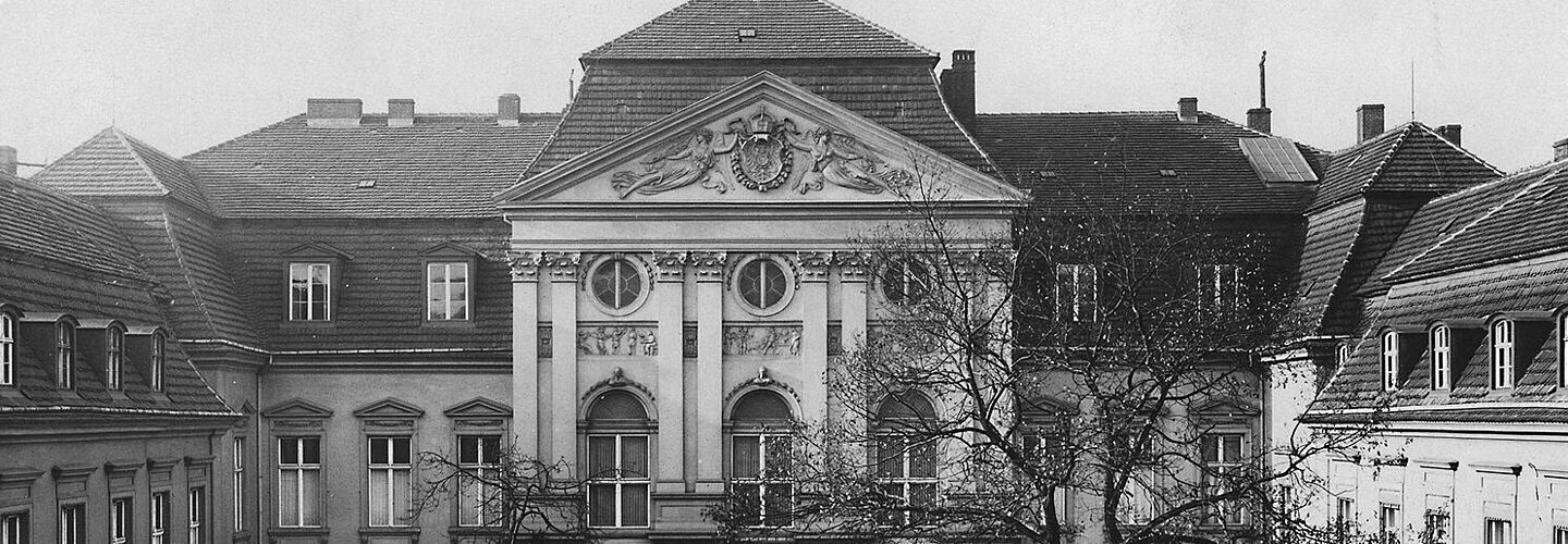 Es handelt sich bei der Schwarz-Weiß-Aufnahme um die Frontansicht des Palais Radziwill in Berlin, das Dienstsitz der Reichskanzlei war. Es befand sich in der Wilhelmstraße 77. Das Gebäude ist in U-Form gebaut. Der Innenhof ist mit einem Rundweg gestaltet, der eine mit Bäumen und Sträuchern bepflanzte Fläche einfasst. Den vorderen Abschluss des Grundstücks bildet ein hoher Metallzaun. Das Bild wurde von einem unbekannten Fotografen aufgenommen.