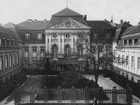 Es handelt sich bei der Schwarz-Weiß-Aufnahme um die Frontansicht des Palais Radziwill in Berlin, das Dienstsitz der Reichskanzlei war. Es befand sich in der Wilhelmstraße 77. Das Gebäude ist in U-Form gebaut. Der Innenhof ist mit einem Rundweg gestaltet, der eine mit Bäumen und Sträuchern bepflanzte Fläche einfasst. Den vorderen Abschluss des Grundstücks bildet ein hoher Metallzaun. Das Bild wurde von einem unbekannten Fotografen aufgenommen.