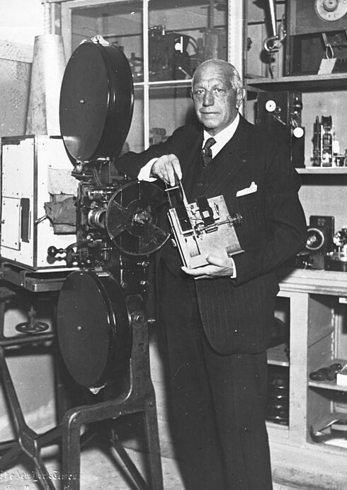 Schwarz-Weiß-Aufnahme von Oskar Messter, der vor einem Kinoprojektor steht und ein technisches Gerät in den Händen hält