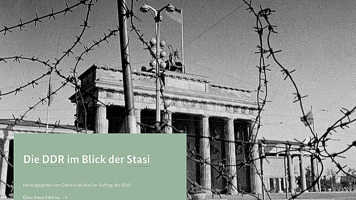 Das Bild zeigt einen Ausschnitt der Startseite der ZAIG-Datenbank 'DDR im Blick der Stasi'. Zu sehen ist ein Schwarz-Weiß-Bild des Brandenburger Tors, das etwas durch Stacheldraht im Vordergrund verdeckt wird. Darüber befindet sich ein mintgrün hinterlegtes Textfeld mit dem Text: ' Die DDR im Blick der Stasi Herausgegeben von Daniela Münkel im Auftrag des BStU'