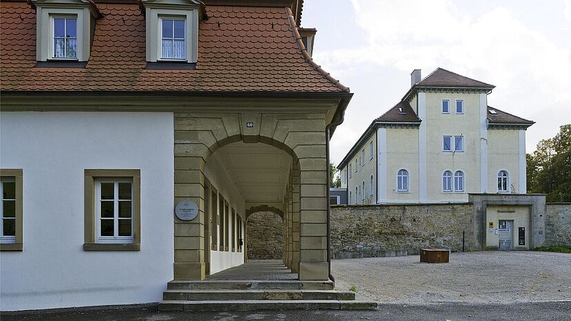 Im Vordergrund steht ein zum Teil weiß verputztes Gebäude. Im Hintergrund ist ein hohes Gebäude hinter einer Mauer zu sehen.